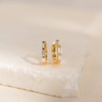 The Kenia Diamond Huggies are small gold earrings with rectangular clear stones, shown upright on a light textured surface—ideal as elegant wedding earrings or chic baguette diamond accessories.