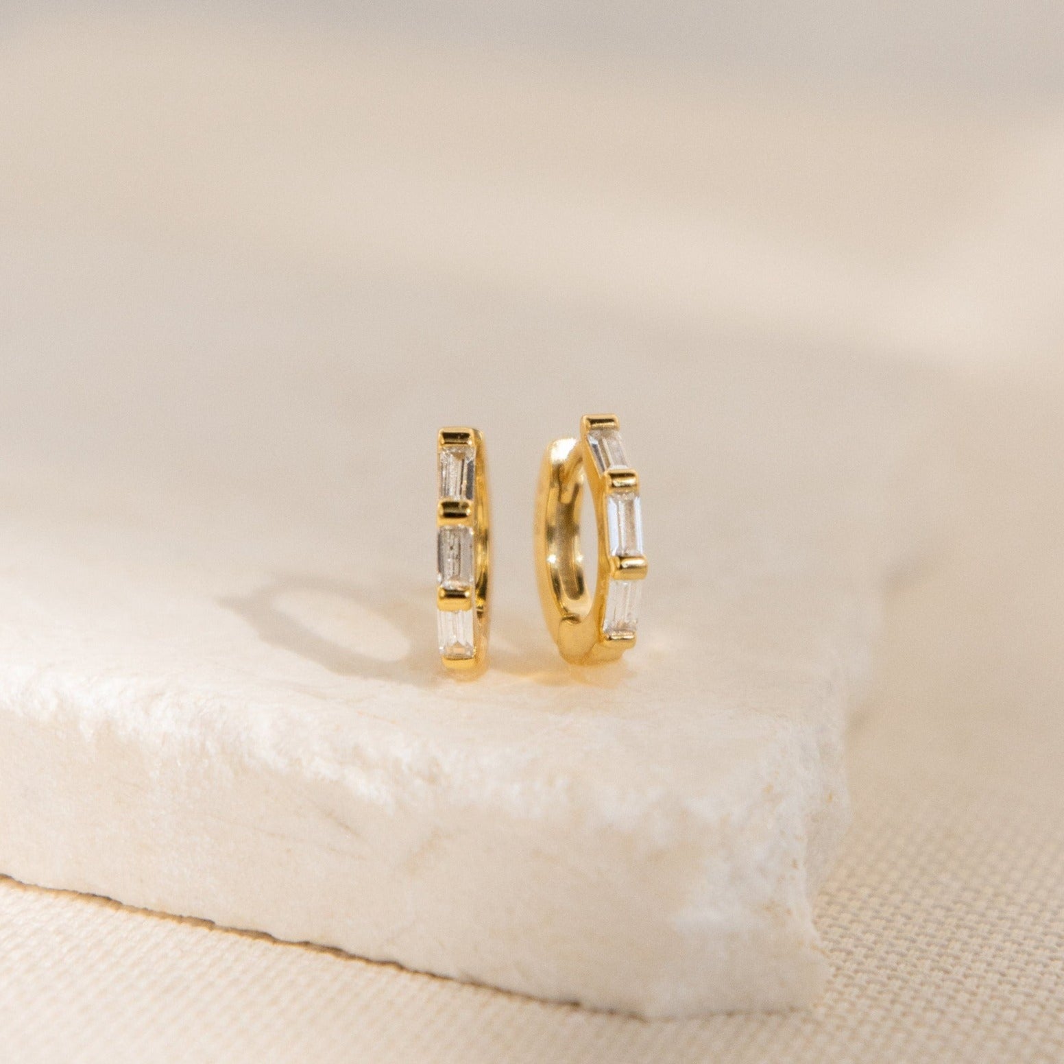 The Kenia Diamond Huggies are small gold earrings with rectangular clear stones, shown upright on a light textured surface—ideal as elegant wedding earrings or chic baguette diamond accessories.