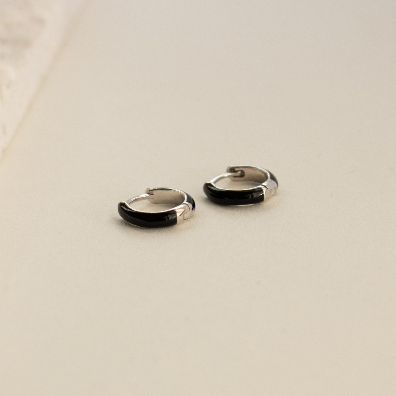 Black Enamel Huggies—small silver and black hoop earrings with sleek onyx enamel—are showcased on a light, neutral surface.