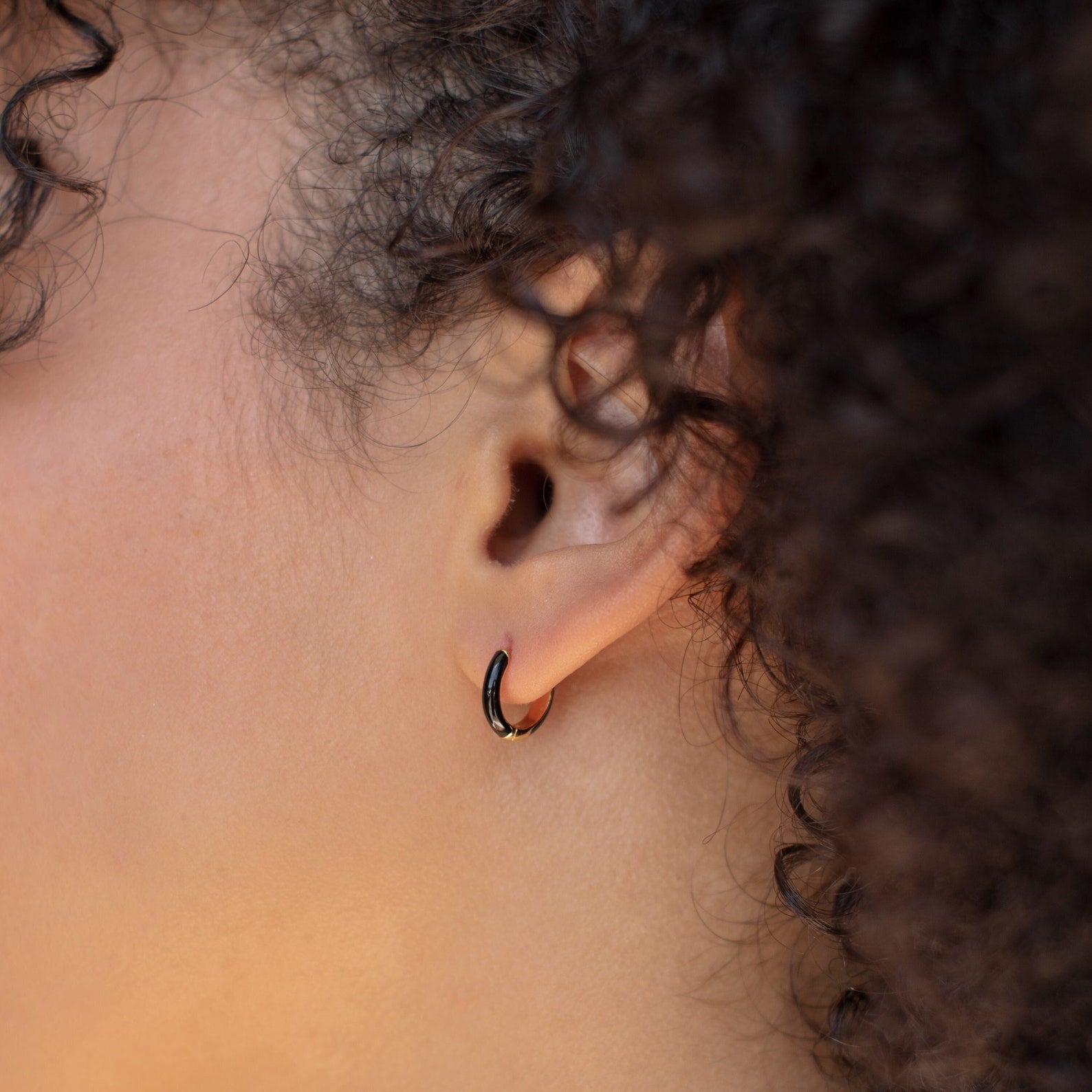 Close-up of an ear adorned with Black Enamel Huggies, a small black hoop earring; curly hair frames the ear.