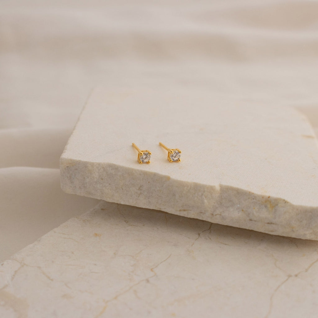 The Booker Classic Diamond Studs are minimalist earrings with small gold settings and clear gemstones, shown on a beige stone slab with a soft, neutral fabric background.