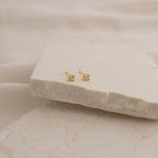 The Booker Classic Diamond Studs are minimalist earrings with small gold settings and clear gemstones, shown on a beige stone slab with a soft, neutral fabric background.