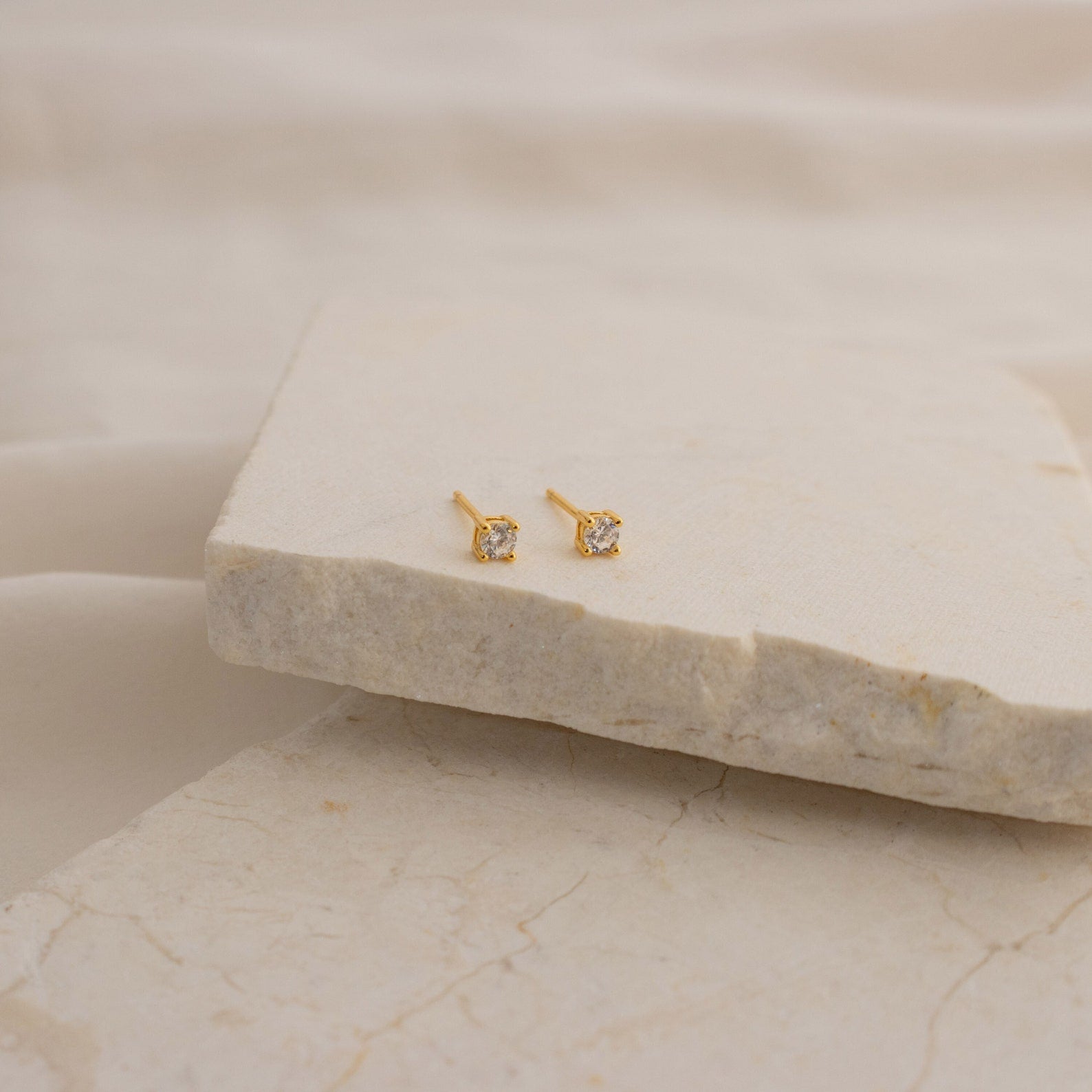 The Booker Classic Diamond Studs are minimalist earrings with small gold settings and clear gemstones, shown on a beige stone slab with a soft, neutral fabric background.