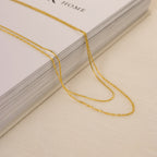 Two gold necklaces laid across a thick hardcover book: one features a sleek snake chain and the other a delicate faceted bead-style chain, both resting on a cream fabric surface.