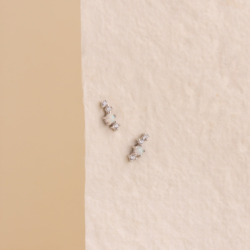 Nile Opal and Diamond Studs—small silver earrings featuring round white stones—are displayed on a textured, off-white background.