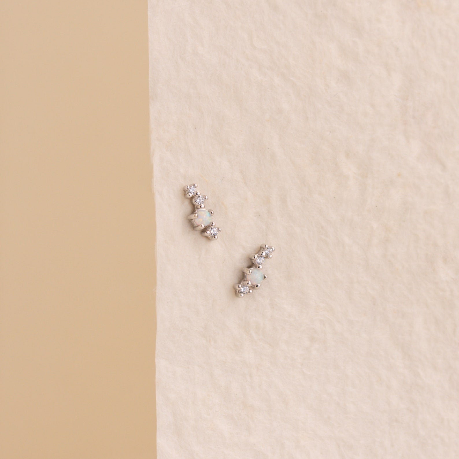 Nile Opal and Diamond Studs—small silver earrings featuring round white stones—are displayed on a textured, off-white background.