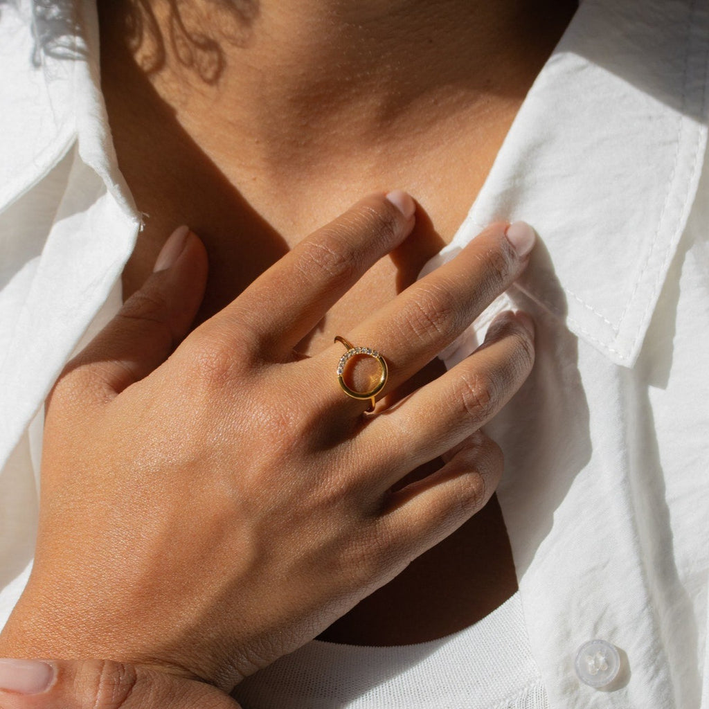 A hand adorned with the Pave Open Circle Ring rests on a chest, wearing a white shirt in sunlight—perfectly highlighting this dainty jewelry.