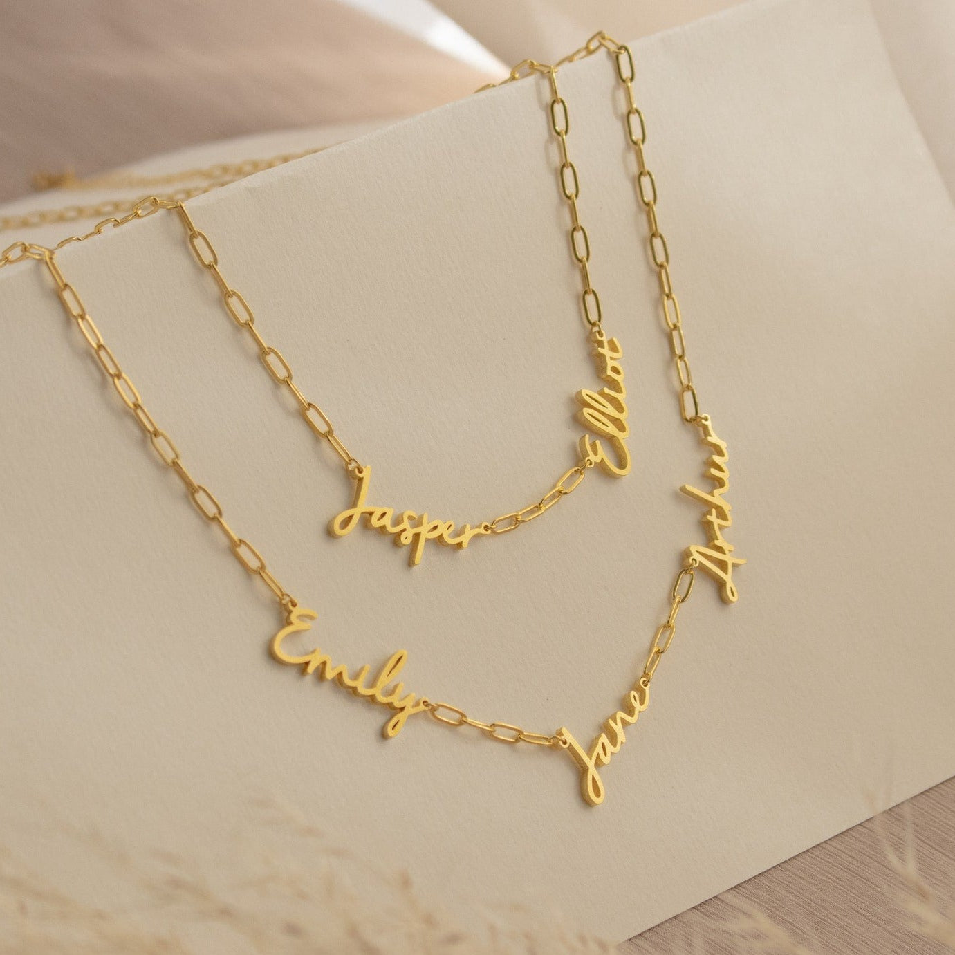 The Nella Multiple Name Necklace in Paperclip Chain features bold link chains with custom cursive pendants—Jasper, Emily, Ellen, Jane, and Aria—for a personalized touch.
