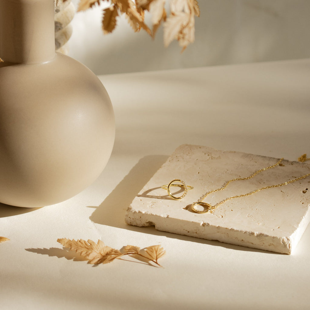 A Pave Open Circle Ring shines on a stone slab alongside gold necklaces and dainty jewelry, styled with a beige vase and dried leaves in natural, soft light.