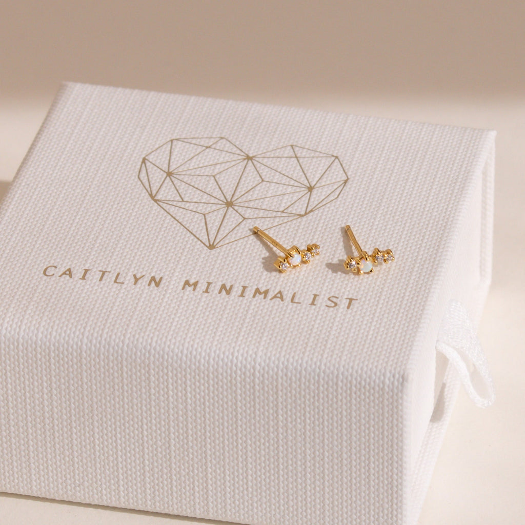 Two Nile Opal and Diamond Studs, inspired by constellations, are displayed on a white textured jewelry box labeled Caitlyn Minimalist.