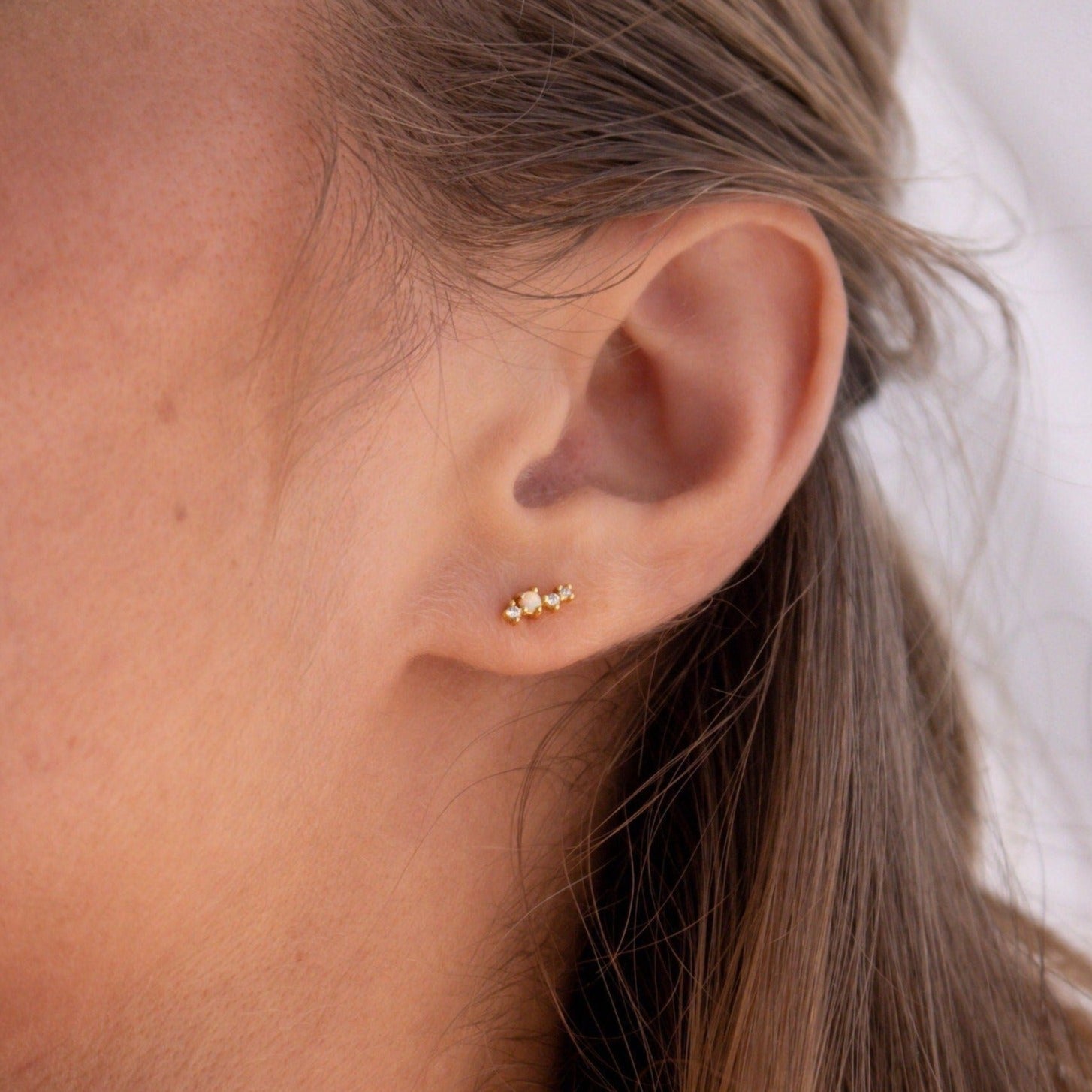 Close-up of a woman's ear wearing Nile Opal and Diamond Studs—small gold floral earrings with constellation-inspired design; her light brown hair is tucked behind her ear.