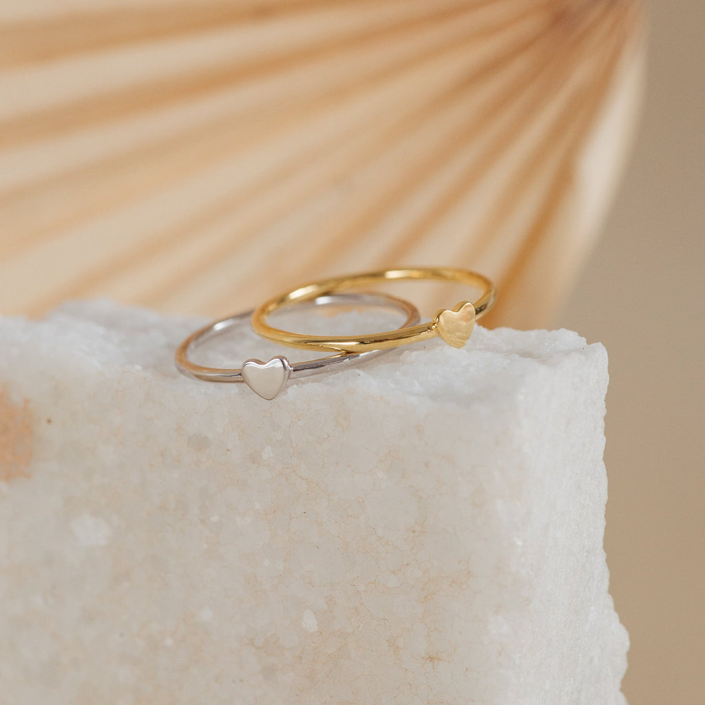 The Nala Tiny Heart Rings—one silver, one gold—each feature a tiny heart and are displayed on a white stone slab. Perfect as promise rings or for everyday wear.