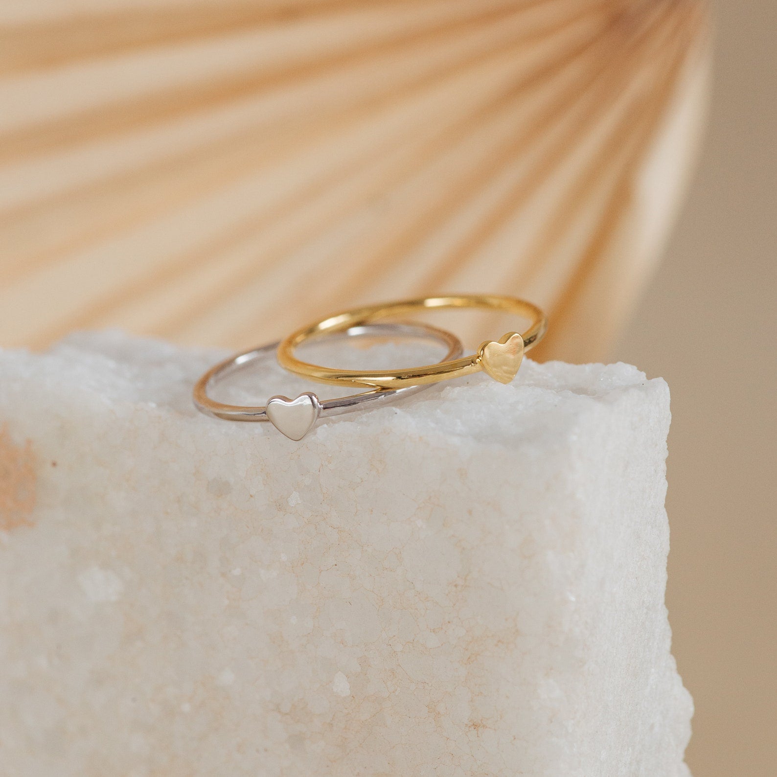 The Nala Tiny Heart Rings—one silver, one gold—each feature a tiny heart and are displayed on a white stone slab. Perfect as promise rings or for everyday wear.