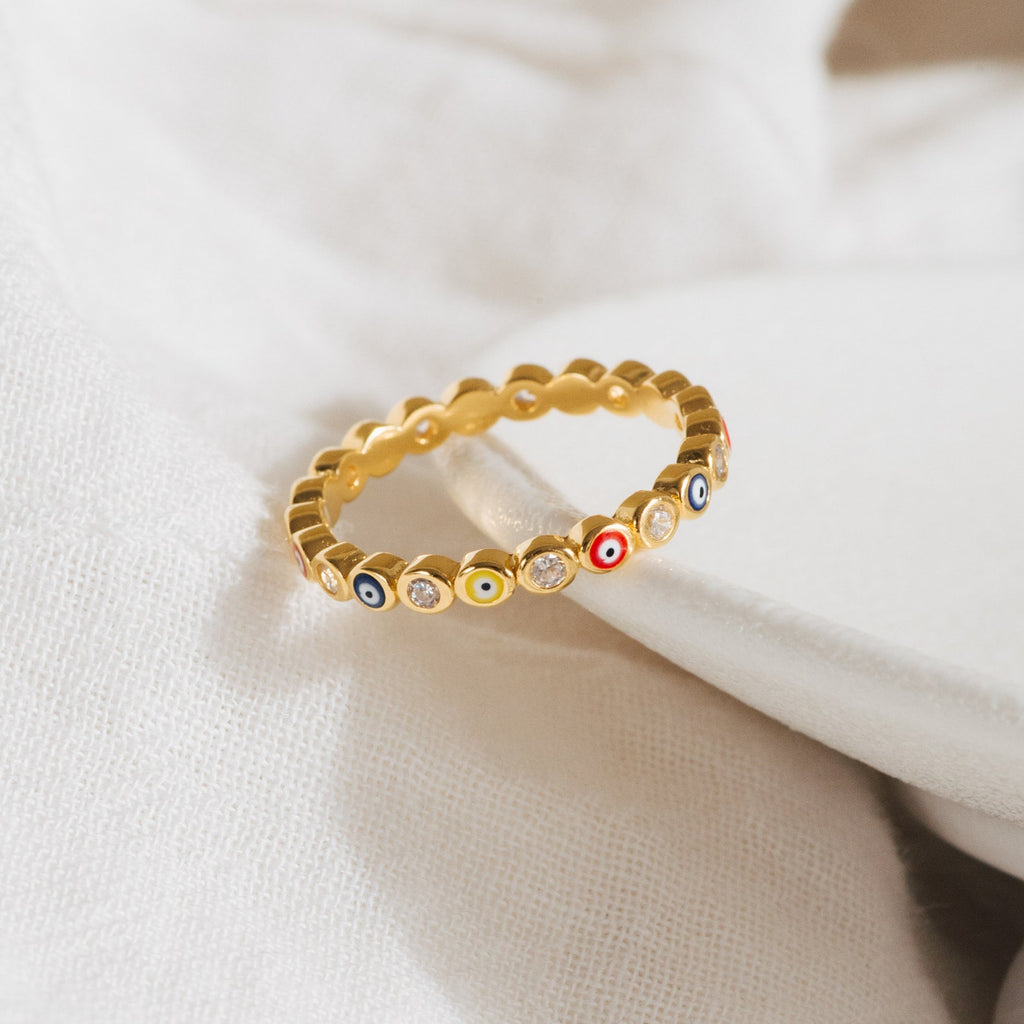 The Rainbow Evil Eye Ring features colorful enamel charms and small crystals, shown on soft white fabric. This vibrant jewelry piece is a stylish, unique lucky charm ring perfect for any occasion.