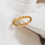 The Rainbow Evil Eye Ring features colorful enamel charms and small crystals, shown on soft white fabric. This vibrant jewelry piece is a stylish, unique lucky charm ring perfect for any occasion.