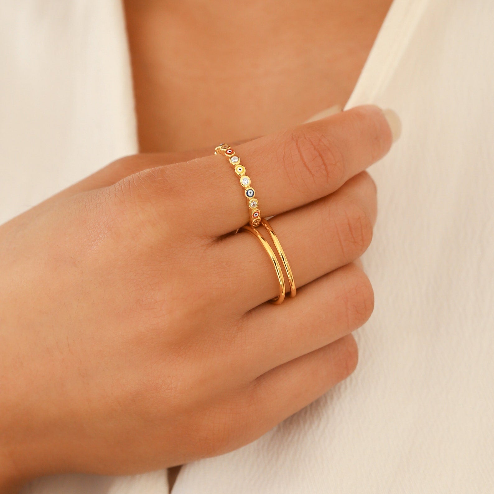 A hand with two elegant gold rings, including the Rainbow Evil Eye Ring adorned with small gems, resting on white fabric.