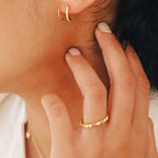 Close-up of a person wearing gold hoop earrings and the Rainbow Evil Eye Ring, featuring colorful stones, as they gently touch their ear.