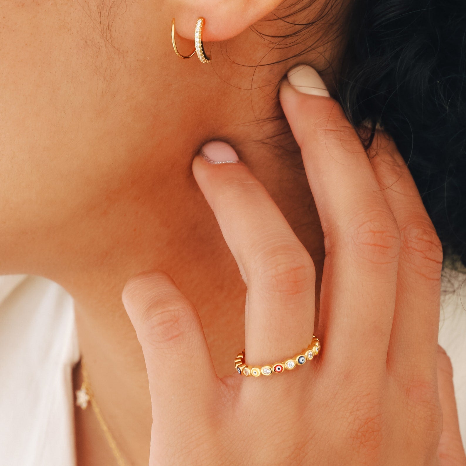 Close-up of a person wearing gold hoop earrings and the Rainbow Evil Eye Ring, featuring colorful stones, as they gently touch their ear.