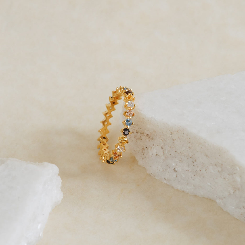 The Rainbow Crystal Ring, a vintage-inspired band with pastel gemstones, shines against a white stone on a beige surface.
