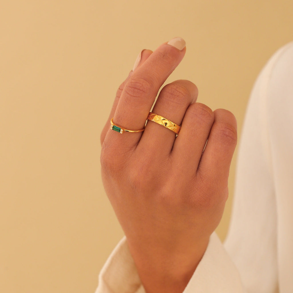 A hand with nude nail polish wears two gold rings: the Empire Emerald Ring and a textured gold band.