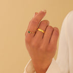 A hand with nude nail polish wears two gold rings: the Empire Emerald Ring and a textured gold band.