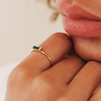 A close-up of a person wearing the Empire Emerald Ring, featuring an elegant emerald set on a gold band, as their fingers gently touch their lips.