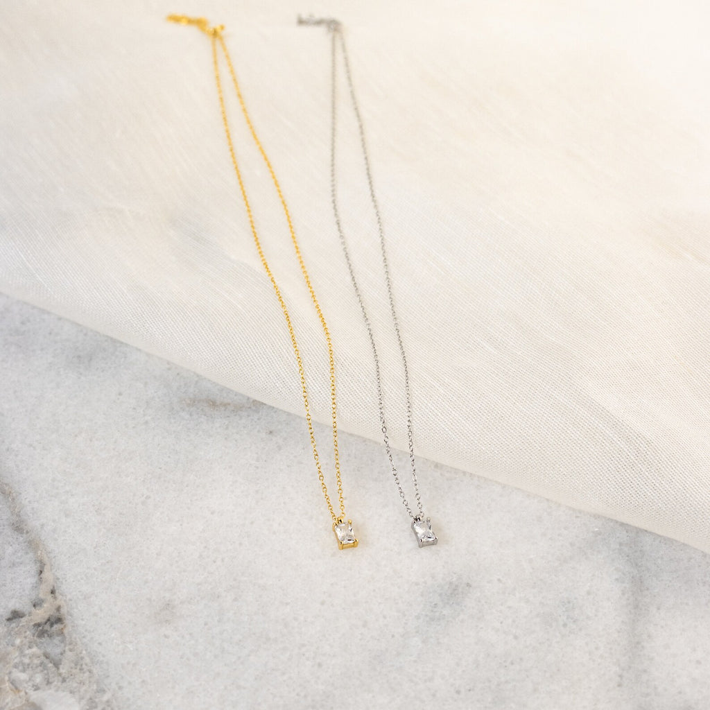 Gold and silver necklaces displayed side by side on a marble surface, each featuring a small rectangular crystal pendant with an emerald cut, styled on a soft cream fabric backdrop for an elegant contrast.