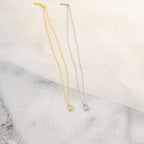 Gold and silver necklaces displayed side by side on a marble surface, each featuring a small rectangular crystal pendant with an emerald cut, styled on a soft cream fabric backdrop for an elegant contrast.