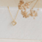 The Forever Pave Heart Necklace features a gold heart pavé design with small crystals, ideal for bridal jewelry, elegantly displayed on white marble with dried flowers.