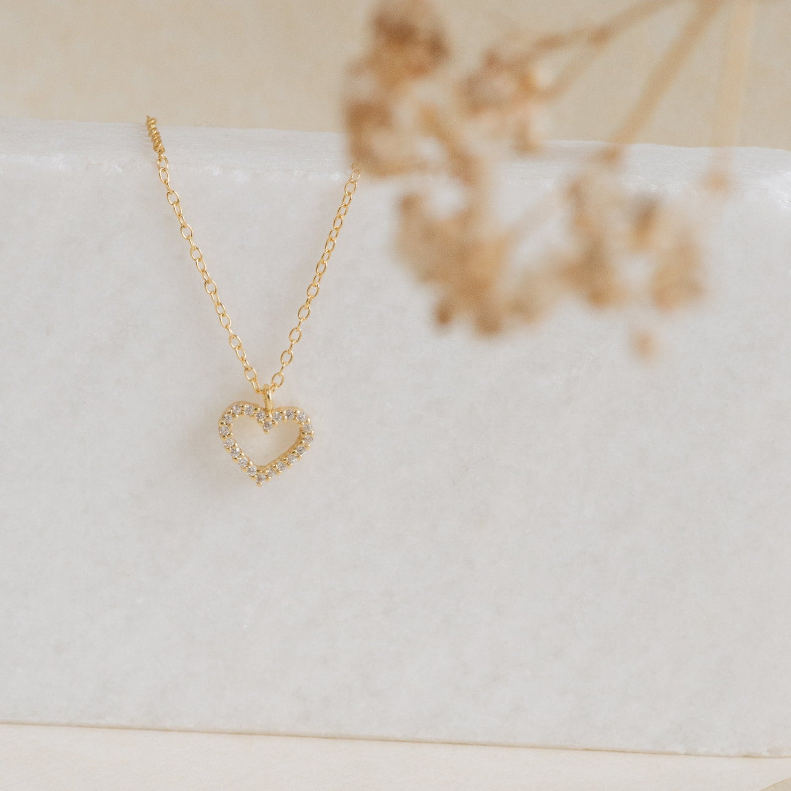 The Forever Pave Heart Necklace features a gold heart pavé design with small crystals, ideal for bridal jewelry, elegantly displayed on white marble with dried flowers.