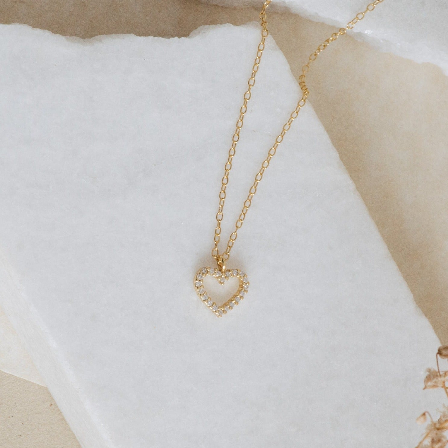 Close-up of a delicate gold necklace featuring an open heart pendant encrusted with small clear crystals, laid flat on a white textured stone surface.