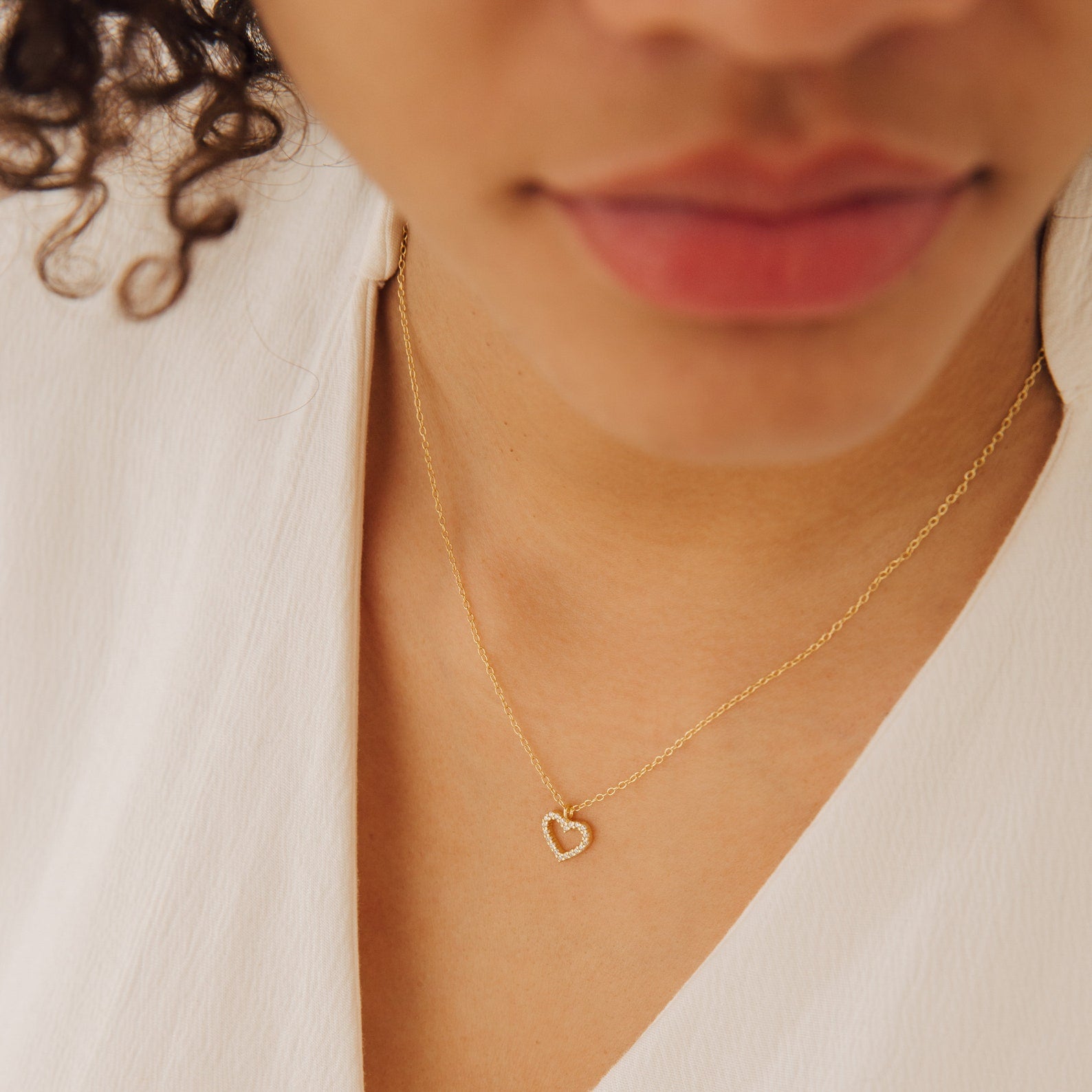 A person wearing the Forever Pave Heart Necklace, featuring a gold chain and small heart-shaped pendant, dressed in a white top.