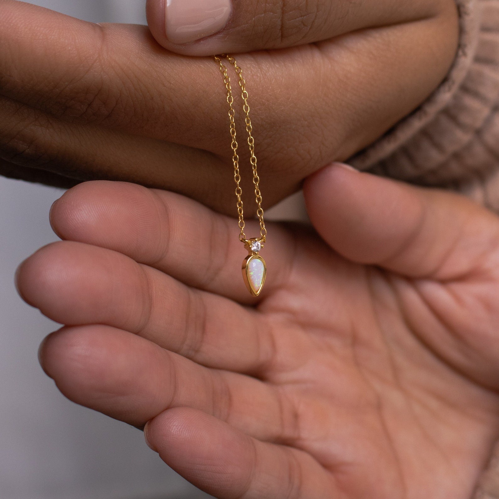 A hand displays the Opal Drop Necklace, featuring a delicate gold chain and an iridescent opal pendant, gently draped over another open palm.