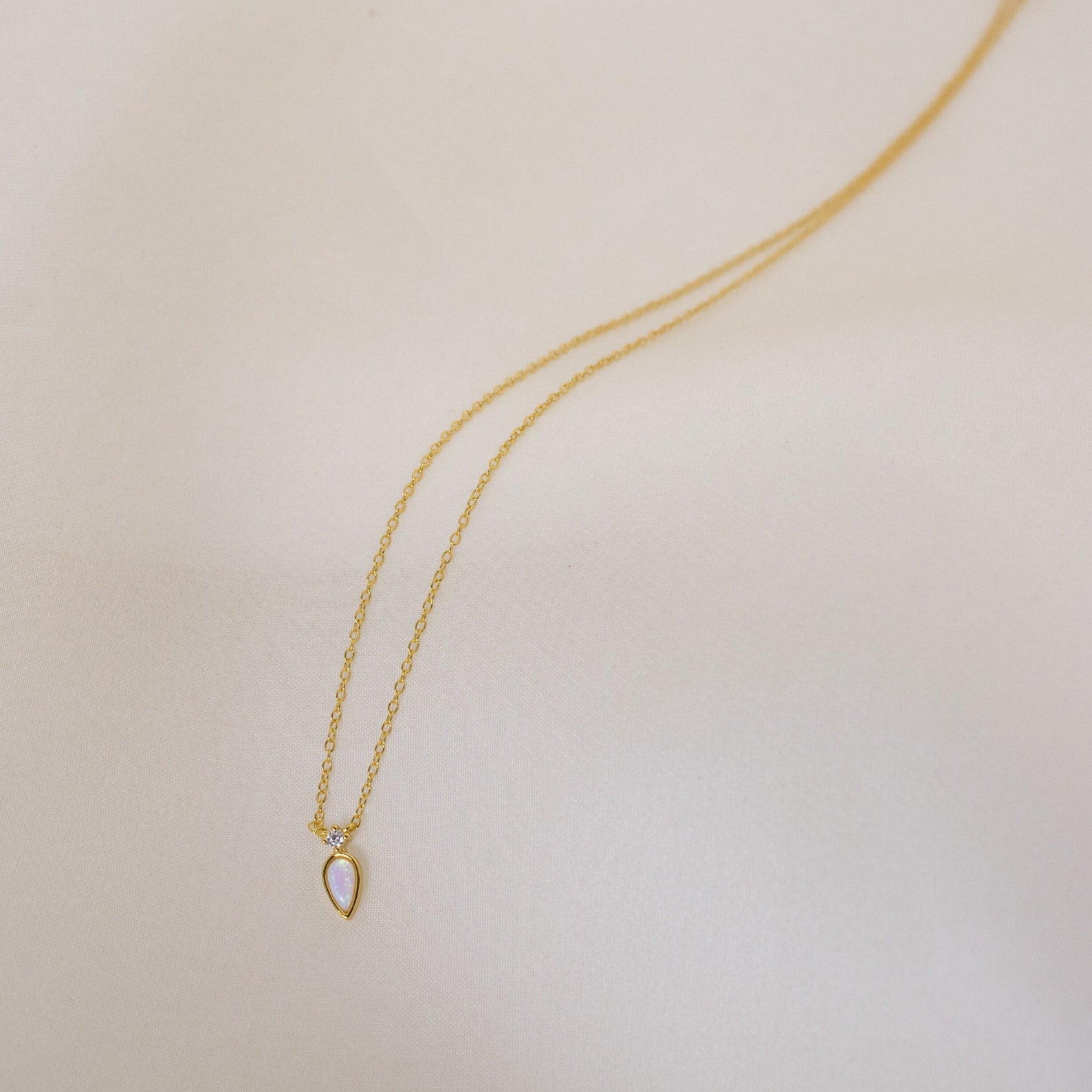 The Opal Drop Necklace features a delicate gold chain with a teardrop-shaped iridescent opal pendant, elegantly displayed on a light, neutral background.