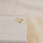 The Booker Tiny Diamond Studs, featuring clear gemstones, rest on a white textured box with a gold geometric heart and the word MINIMALIST, beside a fluffy beige surface.