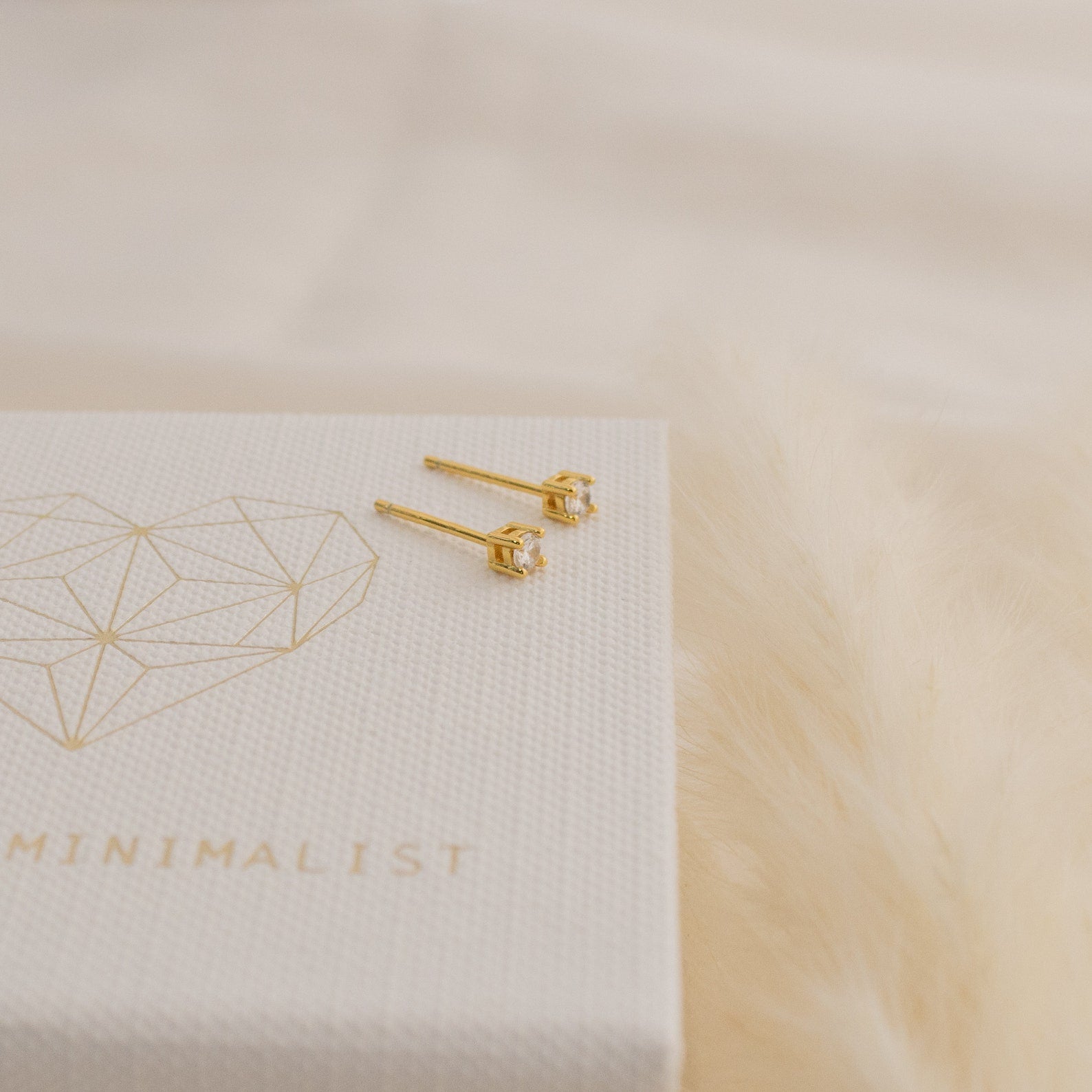 The Booker Tiny Diamond Studs, featuring clear gemstones, rest on a white textured box with a gold geometric heart and the word MINIMALIST, beside a fluffy beige surface.