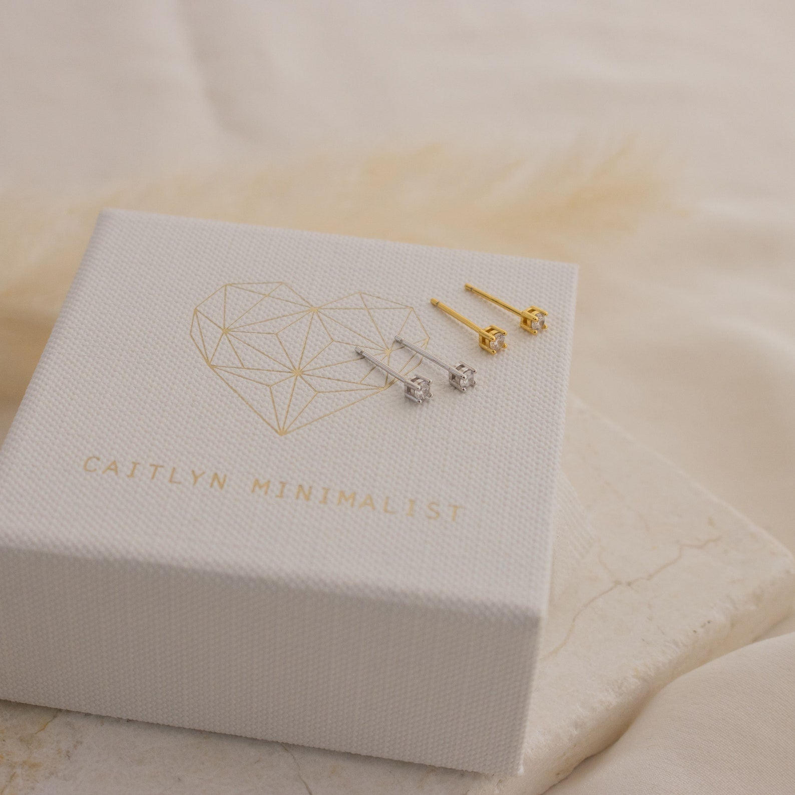 Two pairs of Booker Tiny Diamond Studs—one silver and one gold—are displayed on a white Caitlyn Minimalist jewelry box, perfect for everyday wear.