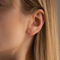 Close-up of a person’s ear with straight blonde hair, wearing the Booker Tiny Diamond Studs—small, round, minimalist earrings set with clear gemstones.