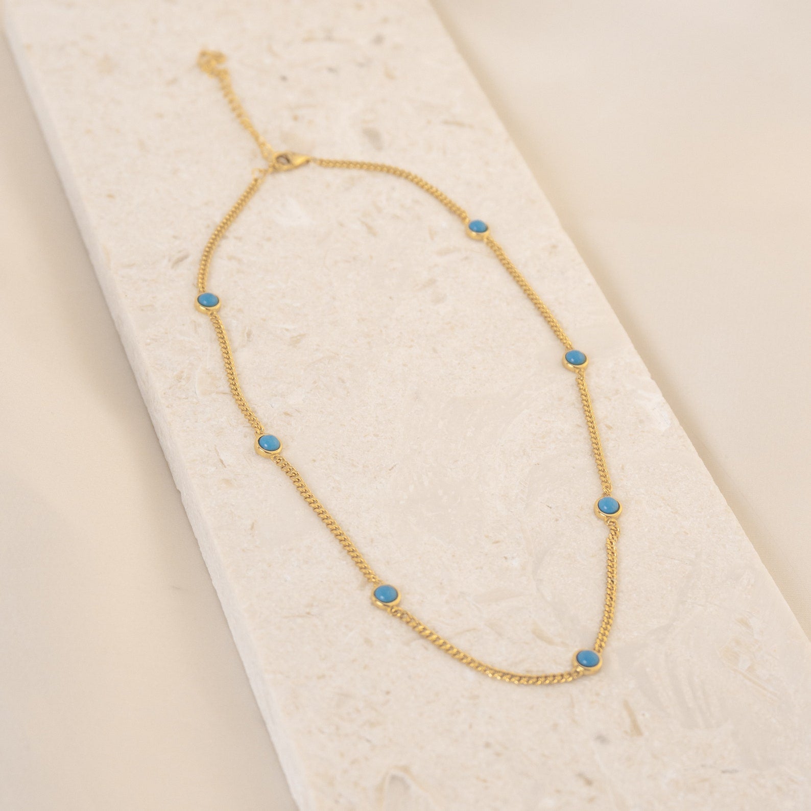 The Santorini Turquoise Choker features a gold chain adorned with small turquoise drops, shown on a light textured background.