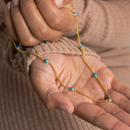 A person wearing a tan ribbed sweater holds the Santorini Turquoise Choker, a gold necklace adorned with delicate turquoise drops.