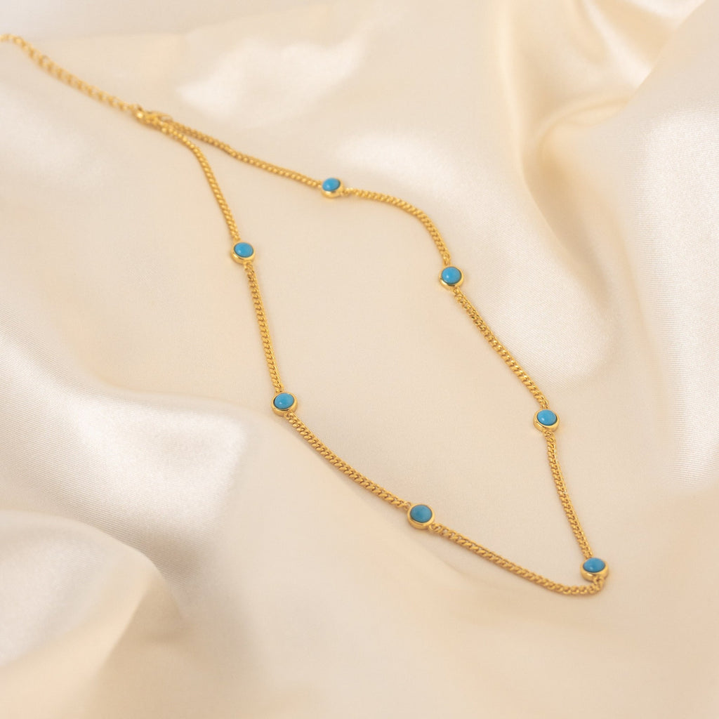 The Santorini Turquoise Choker features a gold chain with evenly spaced turquoise drops, beautifully showcased on smooth cream-colored satin.