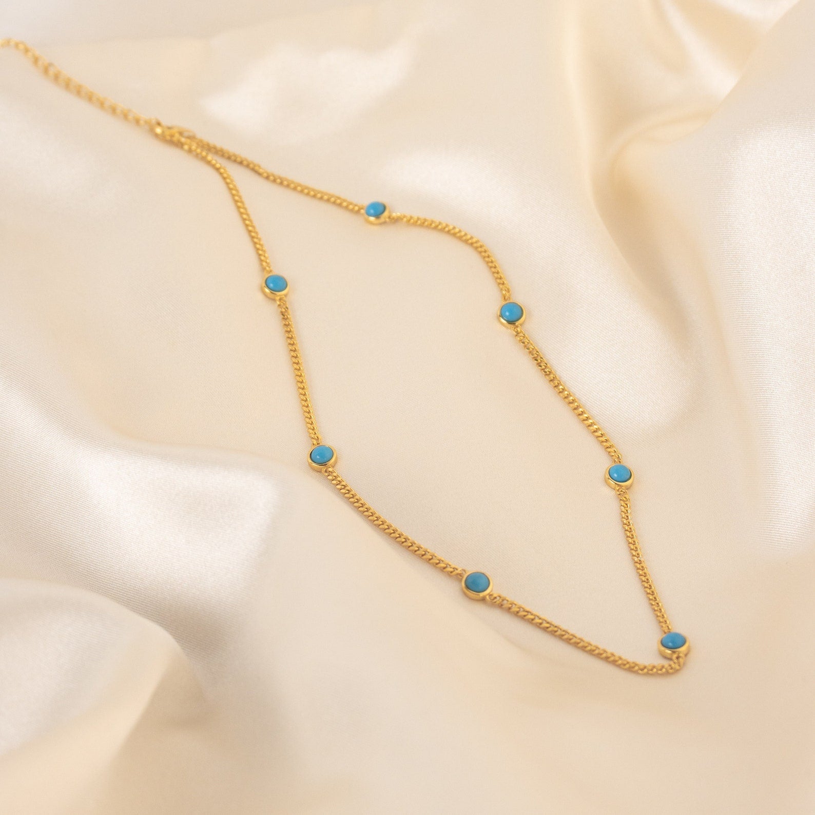 The Santorini Turquoise Choker features a gold chain with evenly spaced turquoise drops, beautifully showcased on smooth cream-colored satin.