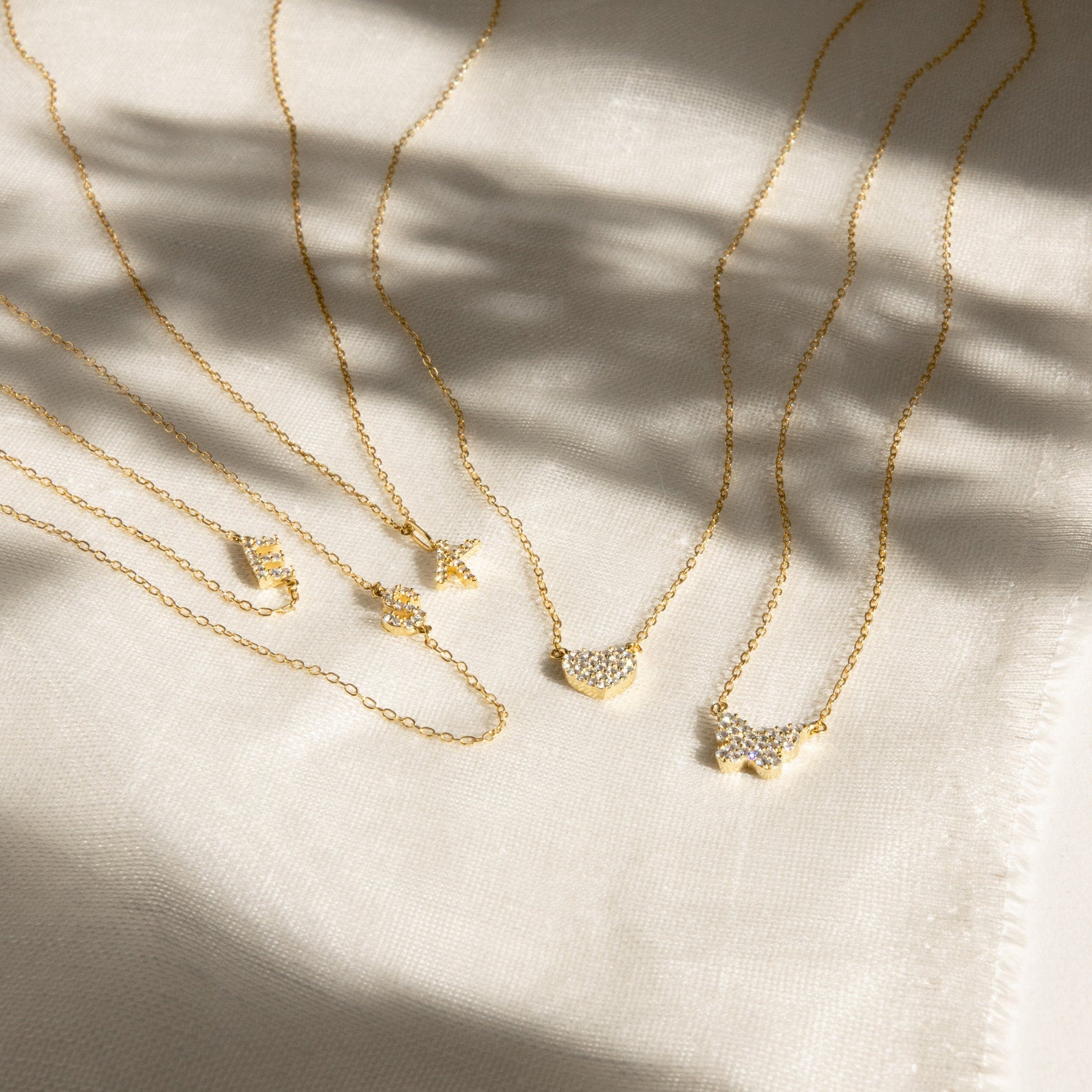 The Pave Initial Necklace, a delicate gold piece with a sparkling pendant, is displayed on light textured fabric—perfect for fans of personalized jewelry.