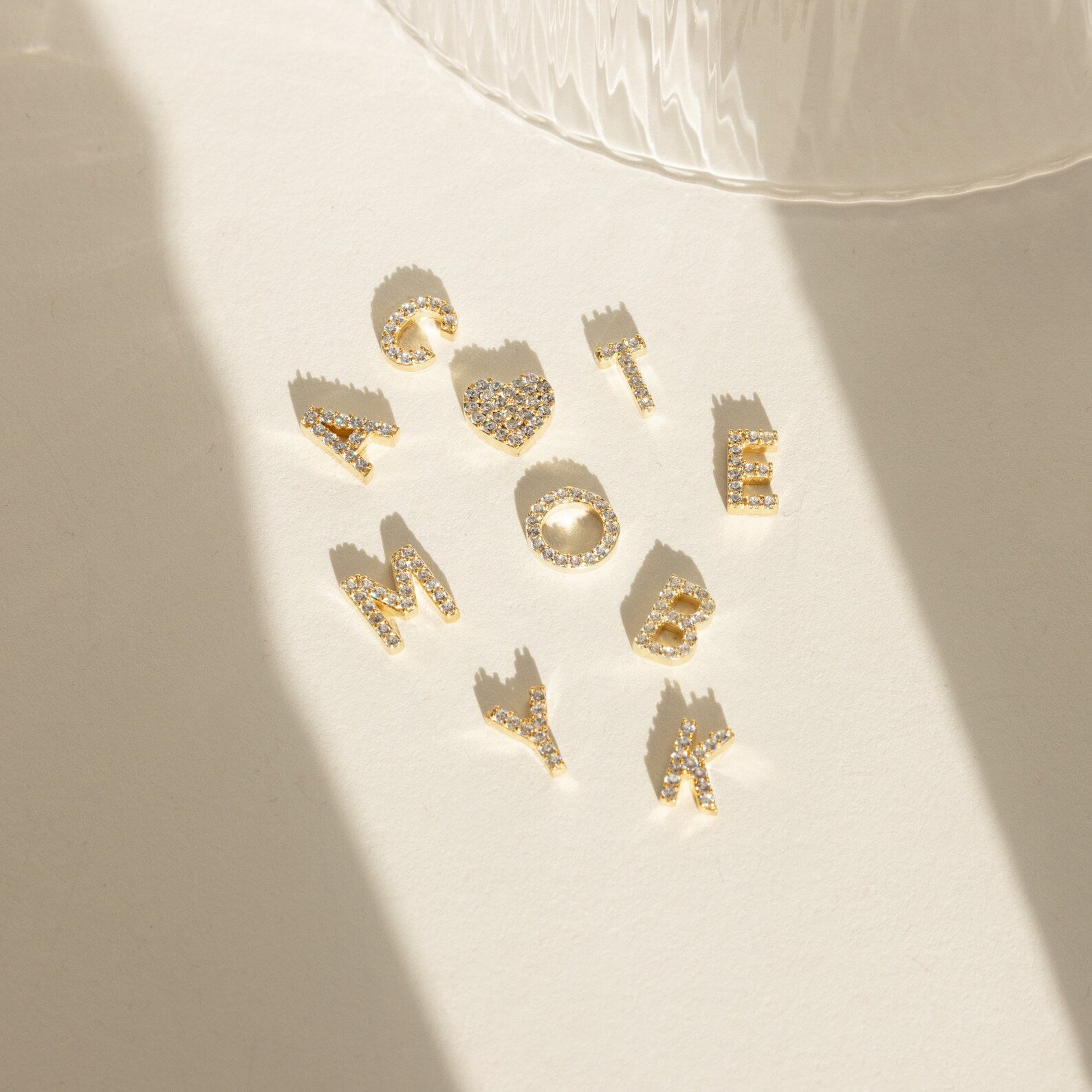 Gold alphabet charms spelling CUTE and AMOYBK on a sunlit surface—our Dangling Pave Initial Necklace is a delicate, sparkling piece, perfect as a necklace or thoughtful gift for women.