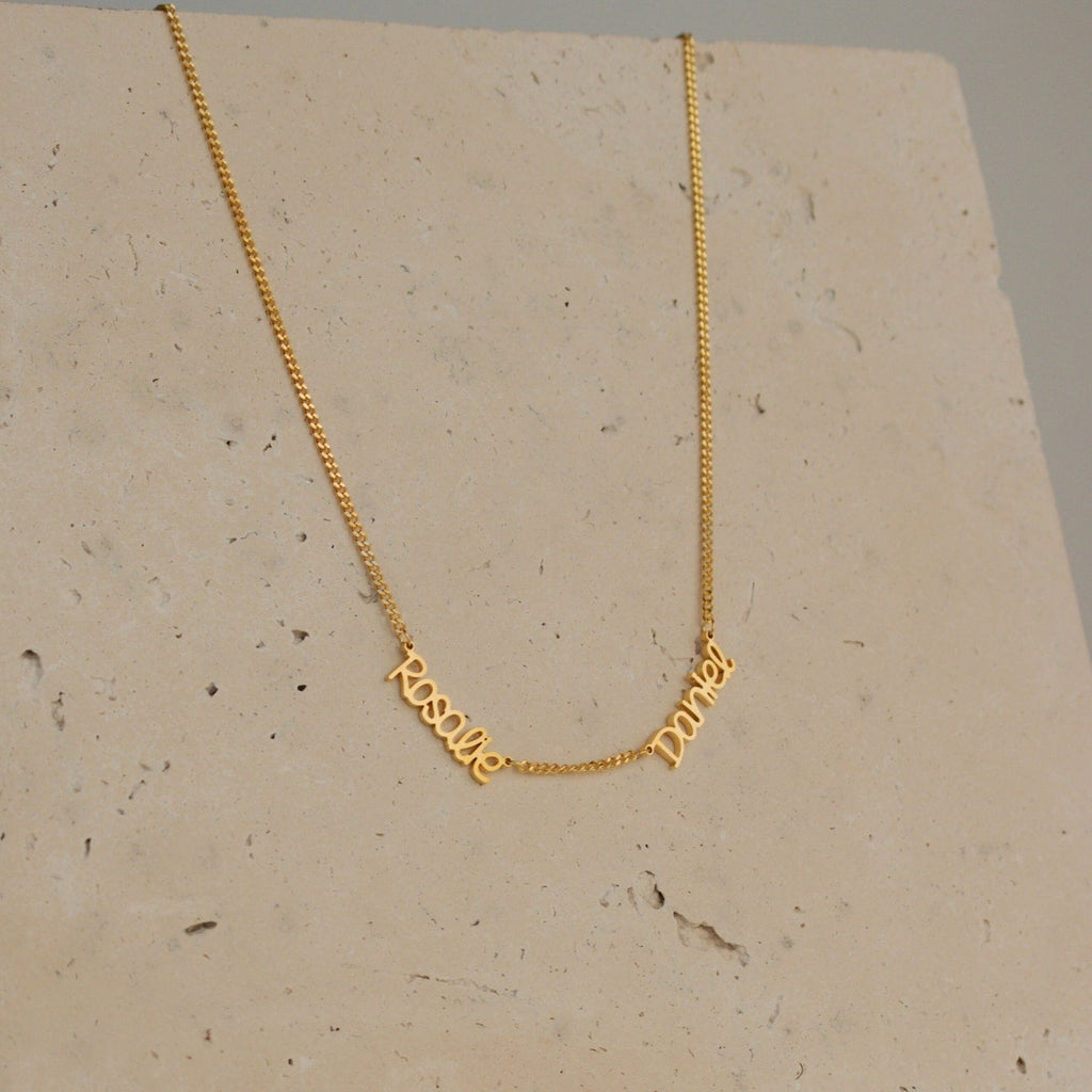 Preppy Multiple Name Necklace in Curb Chain shown on a stone background, featuring the cursive names "Resil" and "Darey" in a youth-inspired font, elegantly connected by a thin curb chain.
