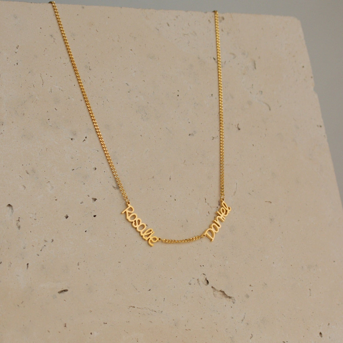Preppy Multiple Name Necklace in Curb Chain shown on a stone background, featuring the cursive names "Resil" and "Darey" in a youth-inspired font, elegantly connected by a thin curb chain.