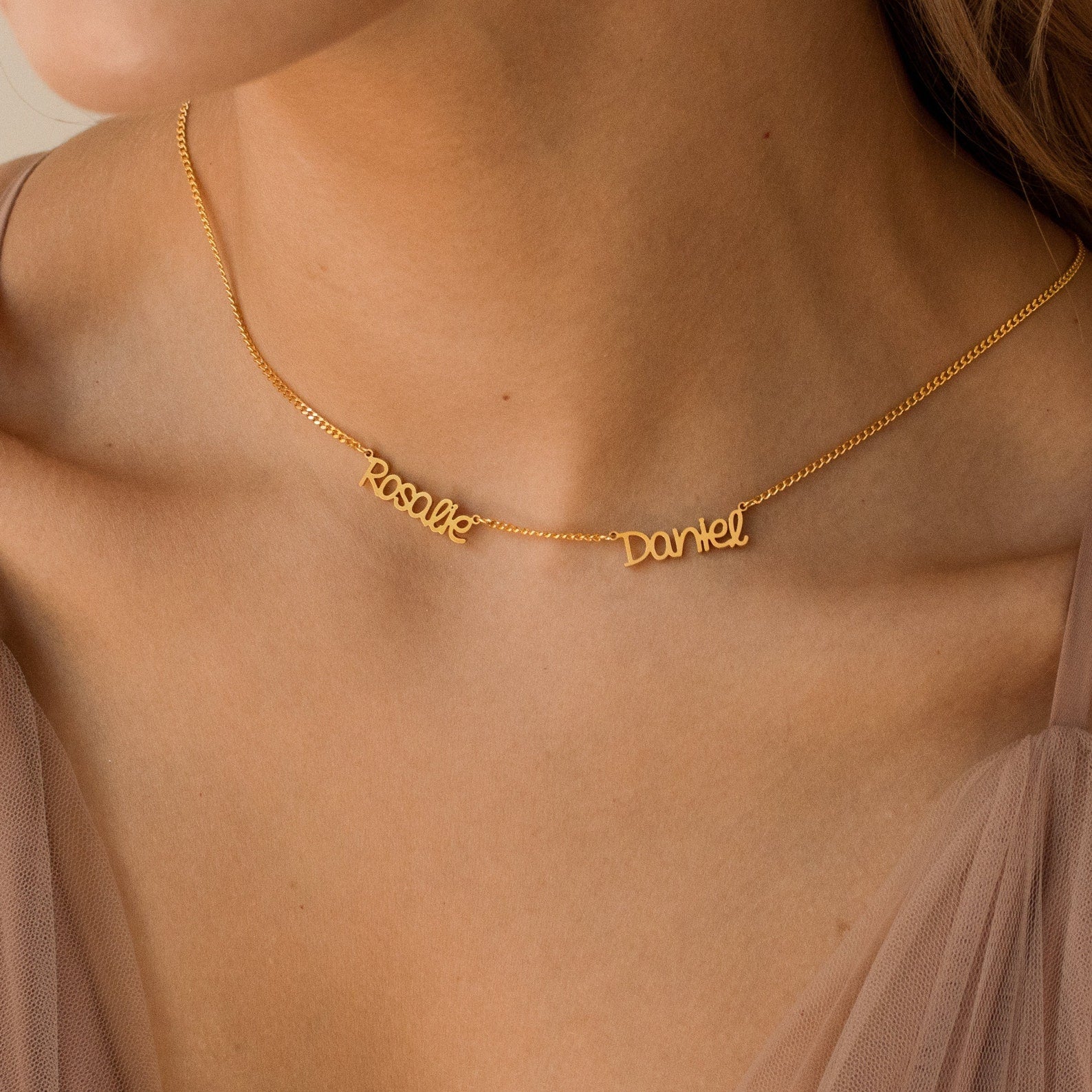 The Preppy Multiple Name Necklace in Curb Chain, featuring names in a youthful font, rests on a woman's neck styled with a beige sheer top and finished with a dainty curb chain.
