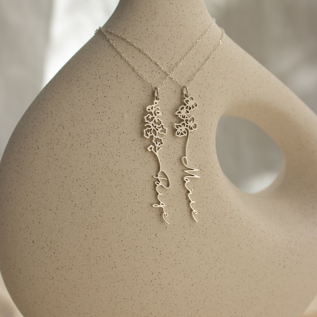 Two delicate silver Flower Name Necklaces with floral designs and personalized name pendants are displayed on a minimalist sculpture, making an ideal gift for anyone who loves unique birth flowers jewelry.