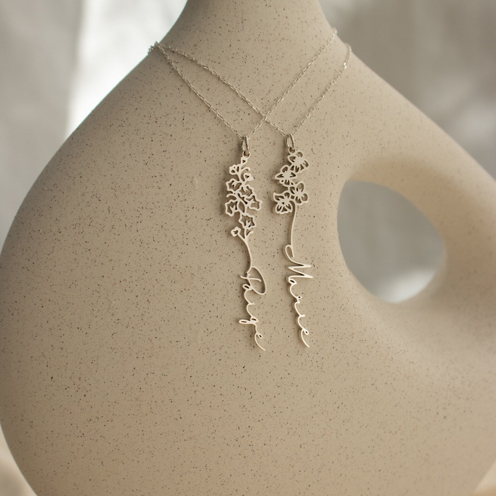 Two delicate silver Flower Name Necklaces with floral designs and personalized name pendants are displayed on a minimalist sculpture, making an ideal gift for anyone who loves unique birth flowers jewelry.