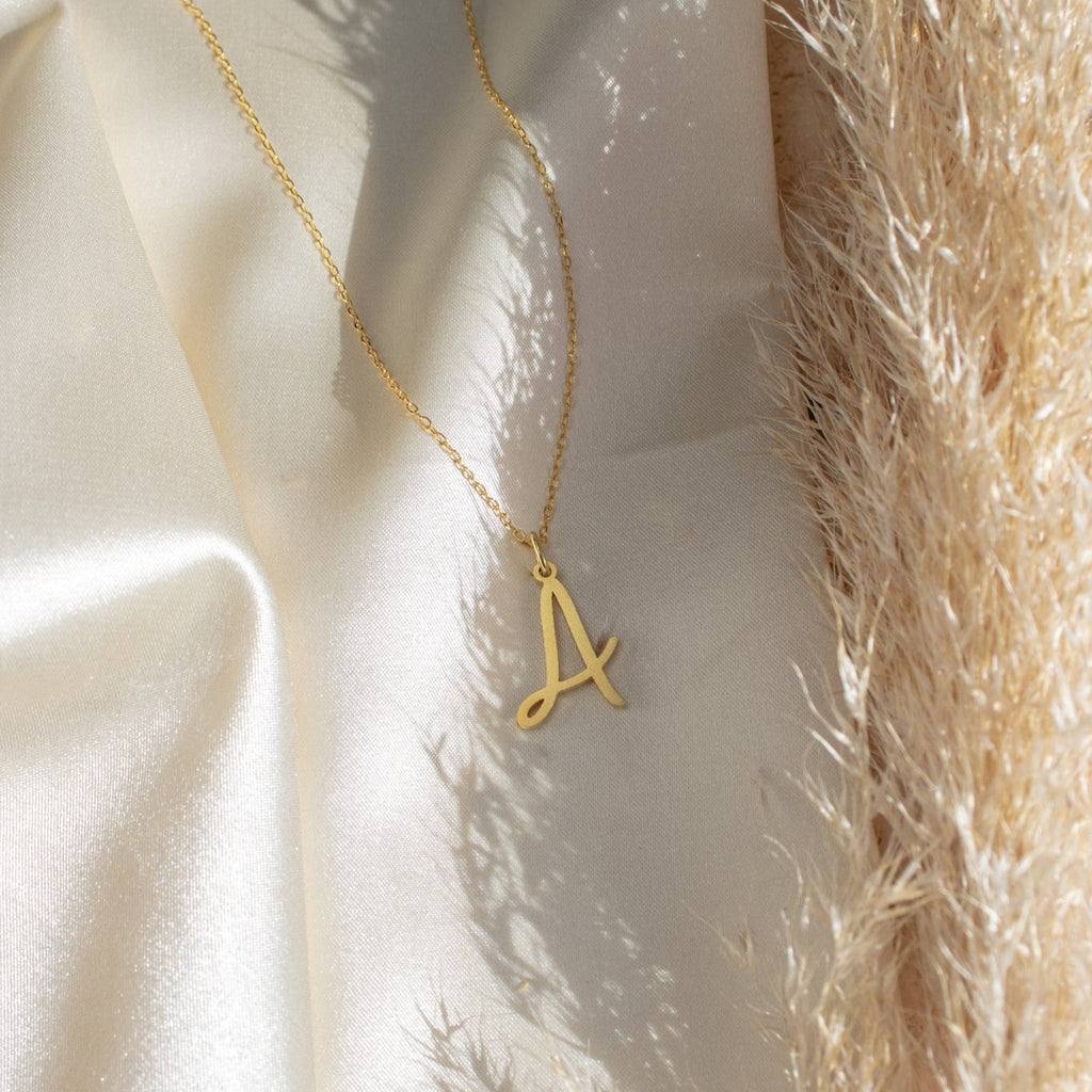 The Nella Letter Necklace with a gold A pendant is displayed on cream satin fabric beside textured dried grass.